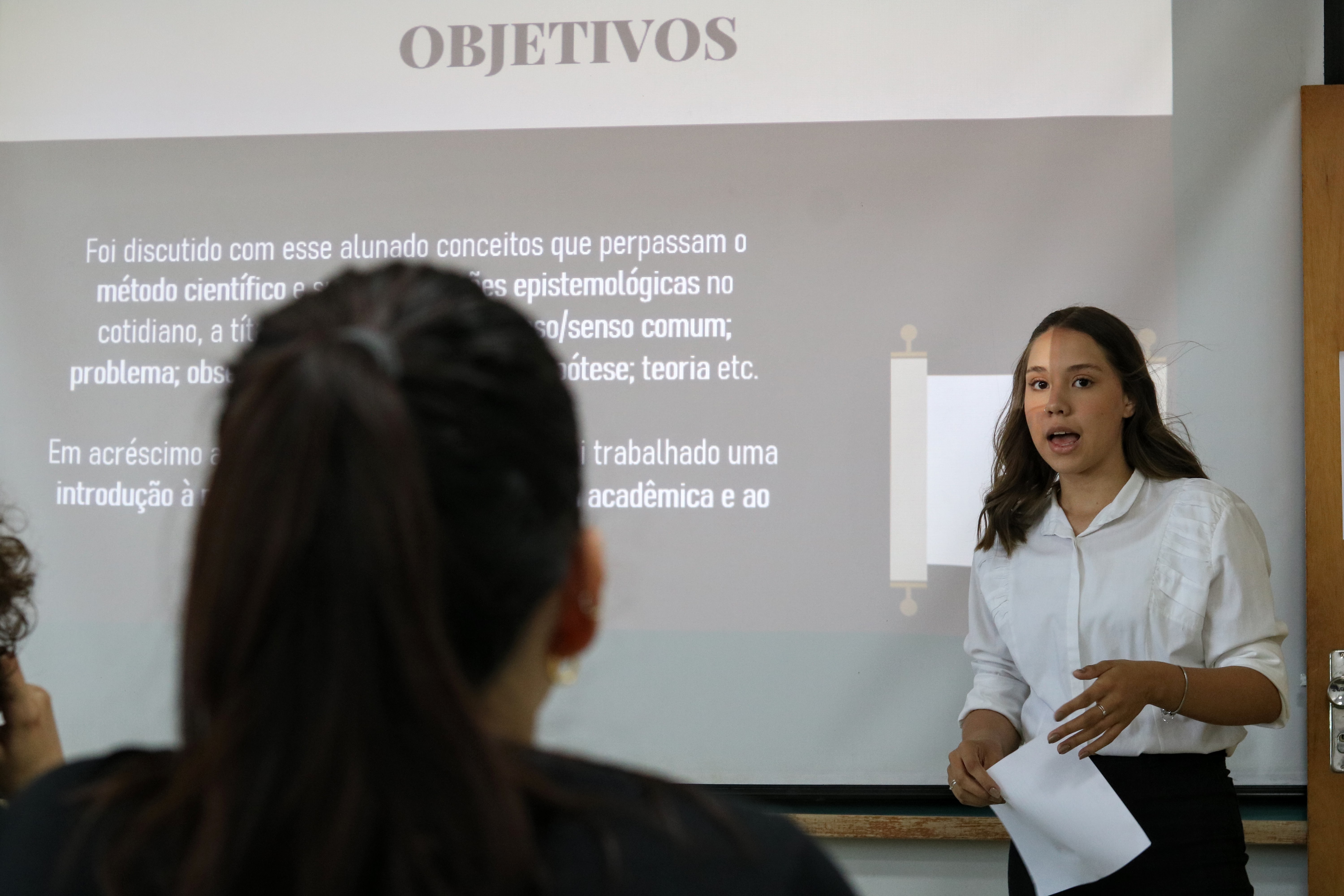 A imagem mostra aluna apresentando pesquisa em uma sala de aula