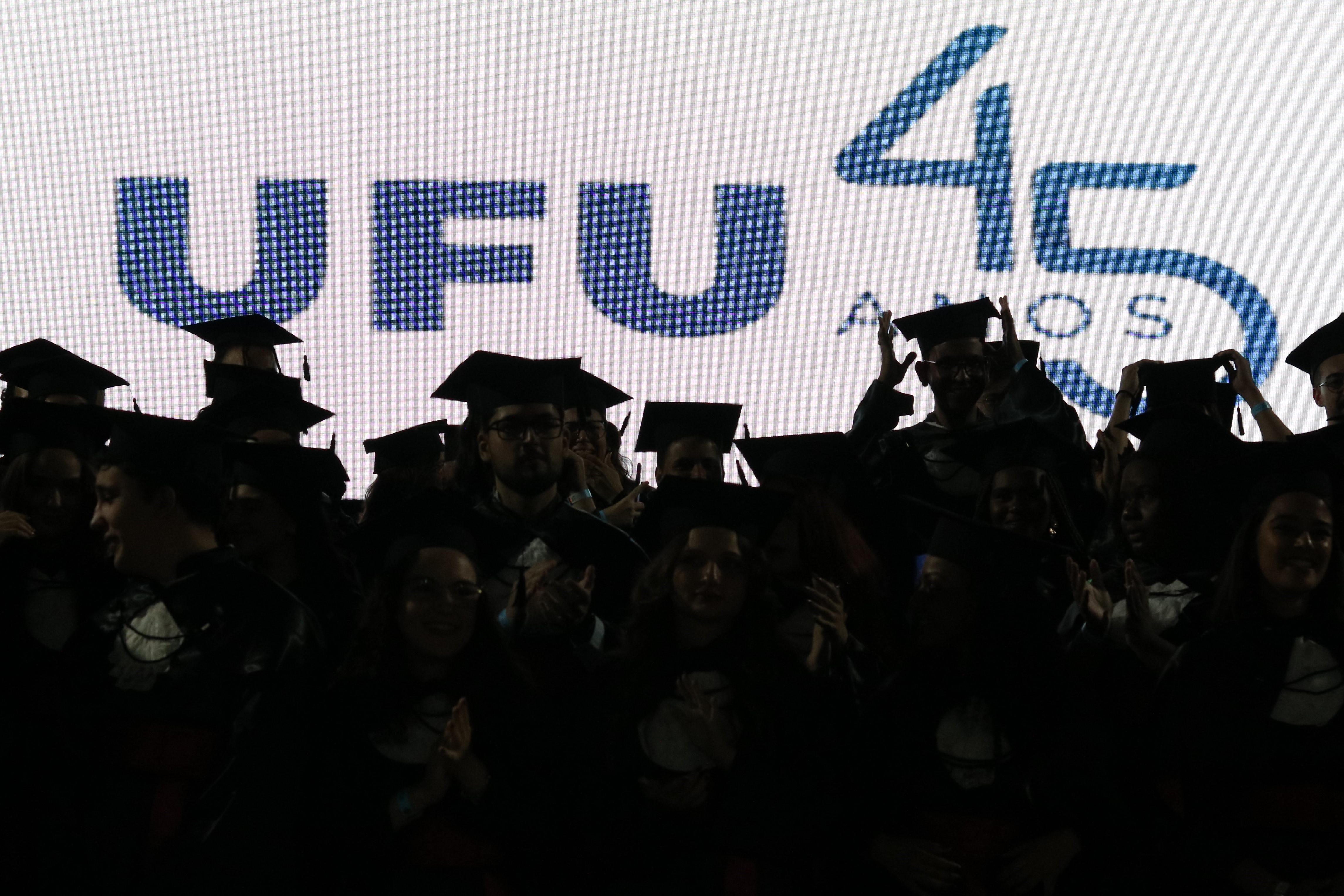 Fundo com logo da UFU 45 anos e mais a frente estão os formandos, formando uma sombra no primeiro plano da imagem.