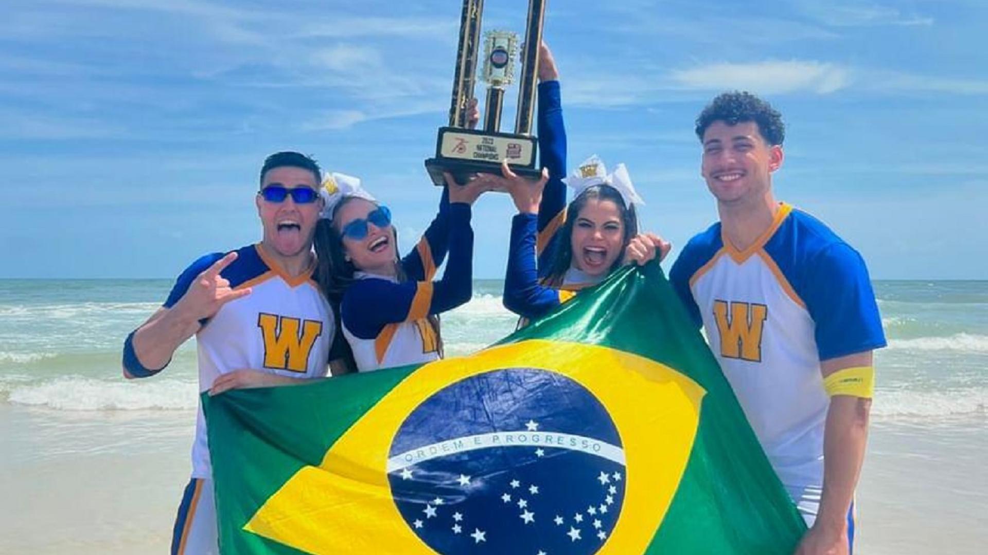 Dois homens e duas mulheres, atletas de cheers, segurando uma bandeira do Brasil e um troféu, na praia