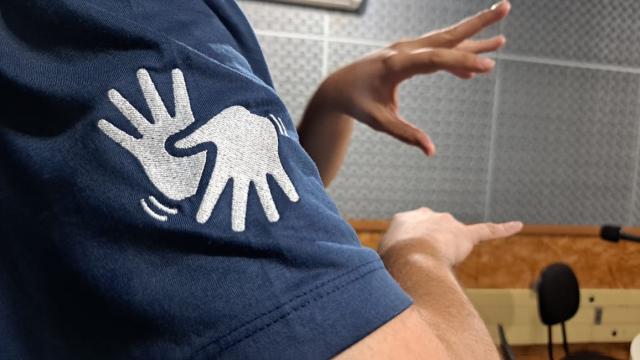 Foto colorida mostra braços de uma pessoa sinalizando em Libras e focando na manga da camiseta que possui o sinal de Libras estampado