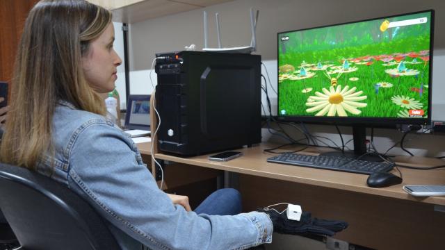 Mulher sentada em frente a uma tela de computador, que exibe imagens do jogo RehaBEElitation em tons verde e amarelo, além de abelhas e flores. Logo abaixo, está a mão e punho da mulher com uma luva vinculada ao game e 'conectada' por pequenos cabos