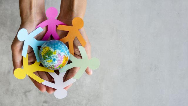 Duas mãos segurando um origami com a imagem de pessoas de cores diferentes, em volta de um globo terrestre 