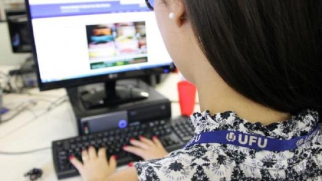 Imagem de mulher de costas, utilizando um crachá da UFU e digitando diante de um monitor de computador