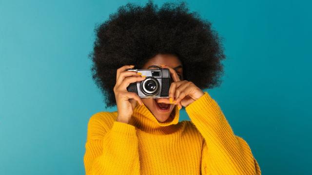 Imagem de moça negra, com cabelo black-power, com rosto escondido atrás de uma câmera fotográfica
