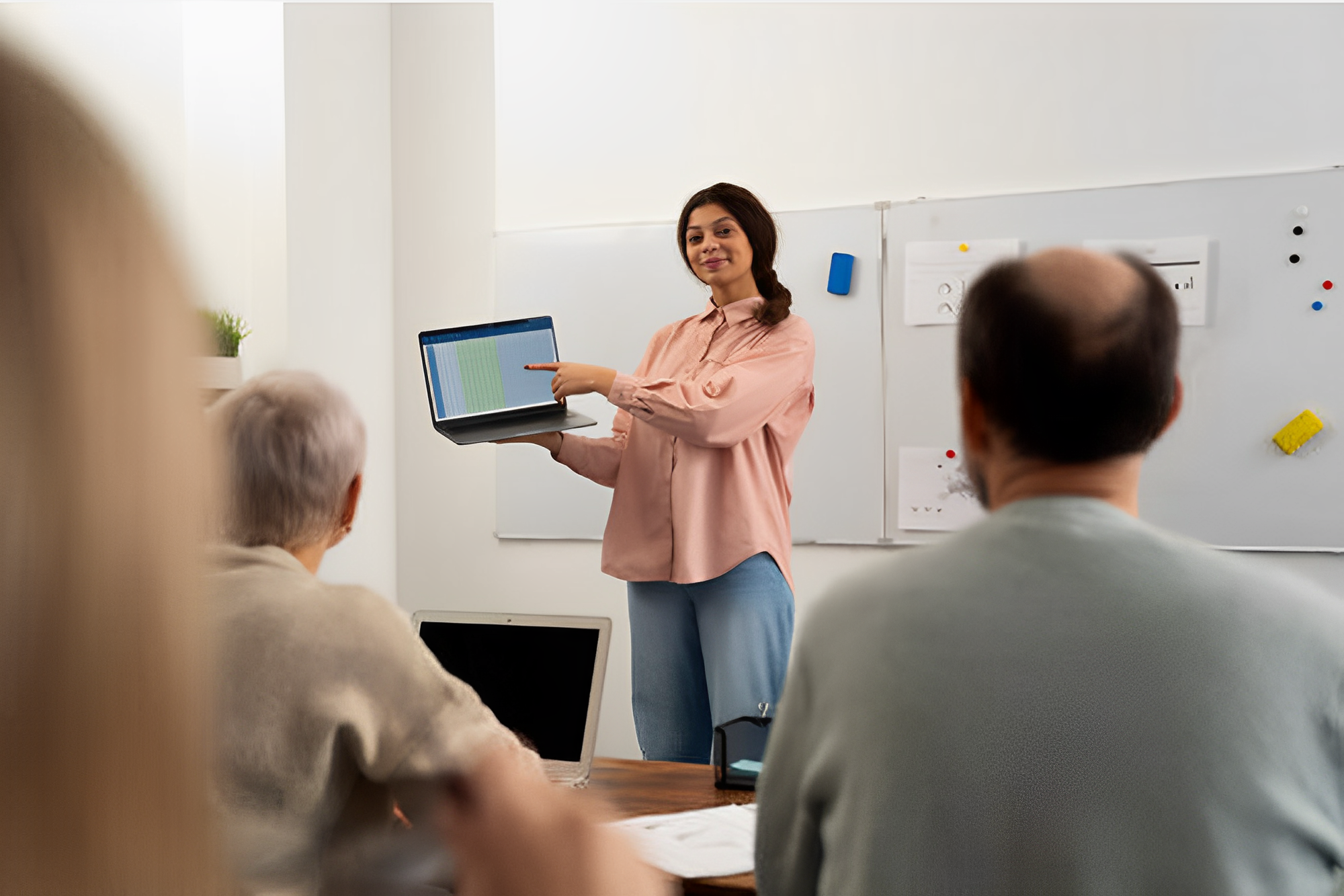 Imagem de uma professora em pé ministrando aula, mostrando a tela de um notebook; em primeiro plano, é possível ver sentados, de costas, uma mulher de cabelo grisalho e um homem calvo