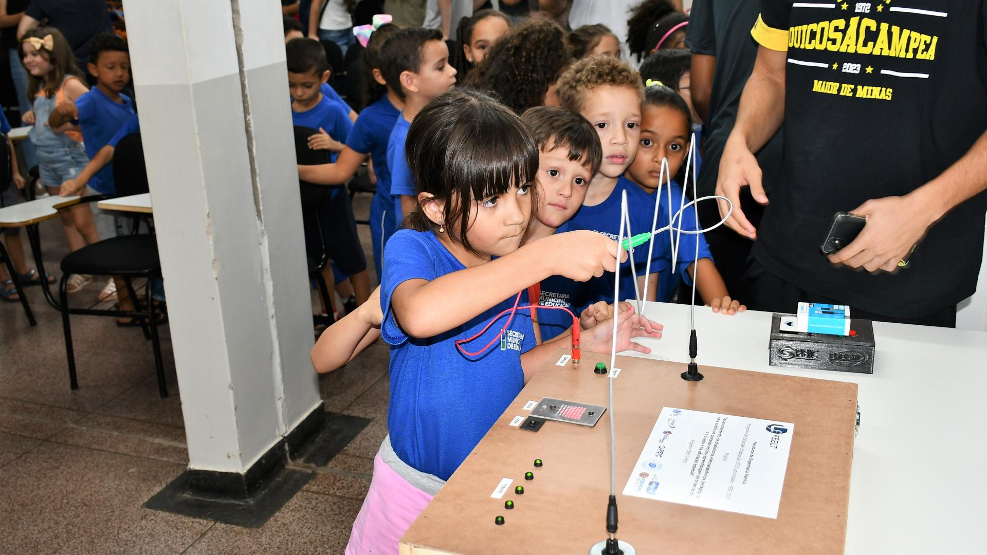 Crianças em fila, aguardando uma delas testar o dispositivo eletroeletrônico, chamado labirinto elétrico, que está sobre a mesa
