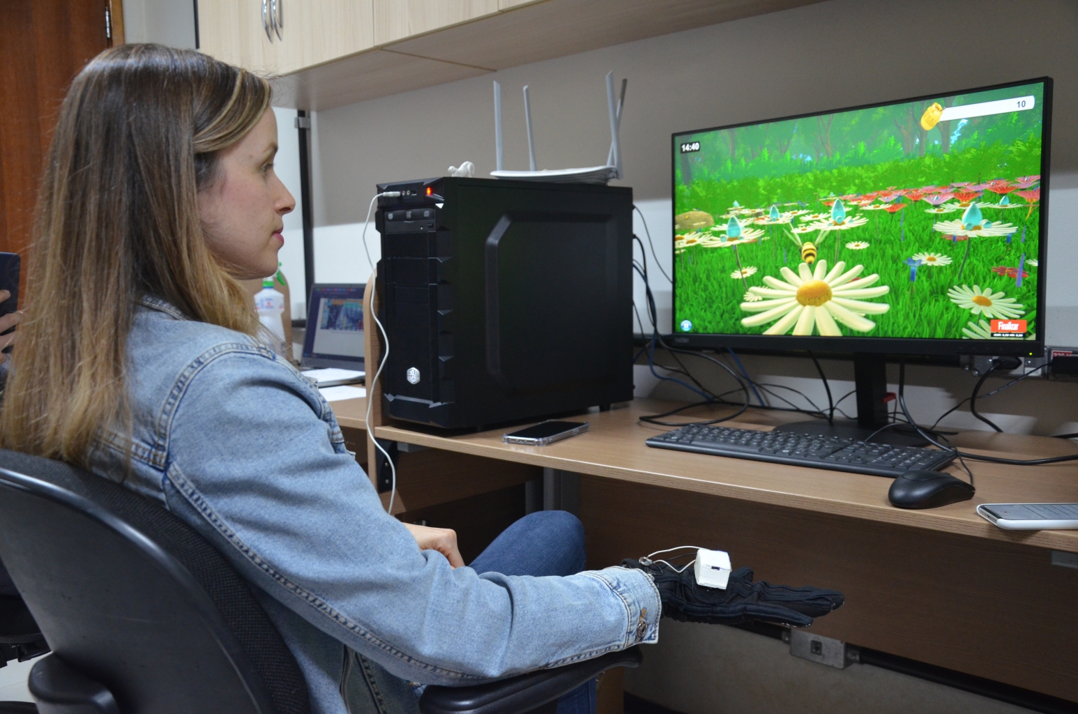 Mulher sentada em frente a uma tela de computador, que exibe imagens do jogo RehaBEElitation em tons verde e amarelo, além de abelhas e flores. Logo abaixo, está a mão e punho da mulher com uma luva vinculada ao game e 'conectada' por pequenos cabos