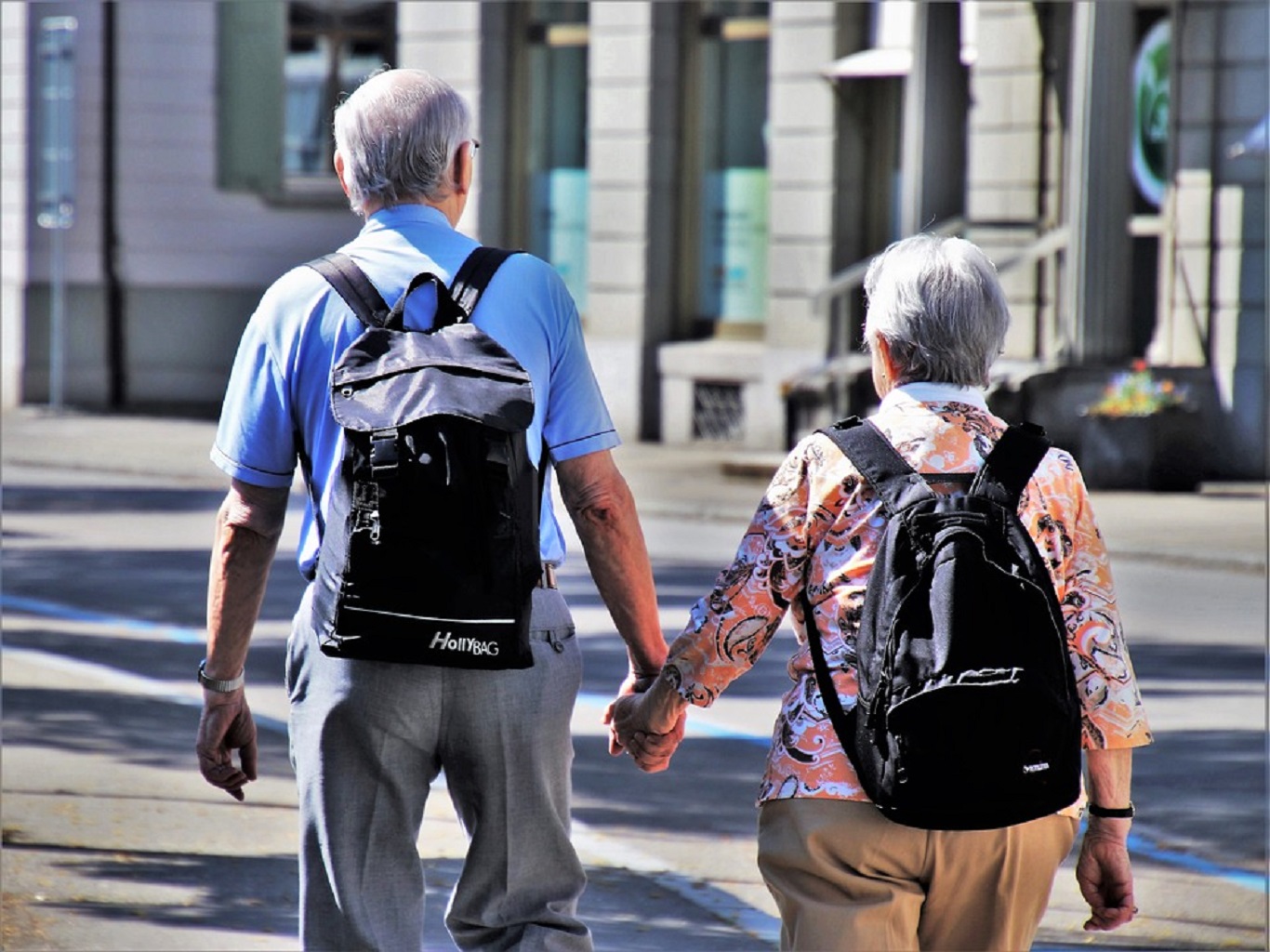 Imagem de um homem e uma mulher de costas, ambos com cabelos grisalhos e mochilas na costas, caminhando de mãos dadas