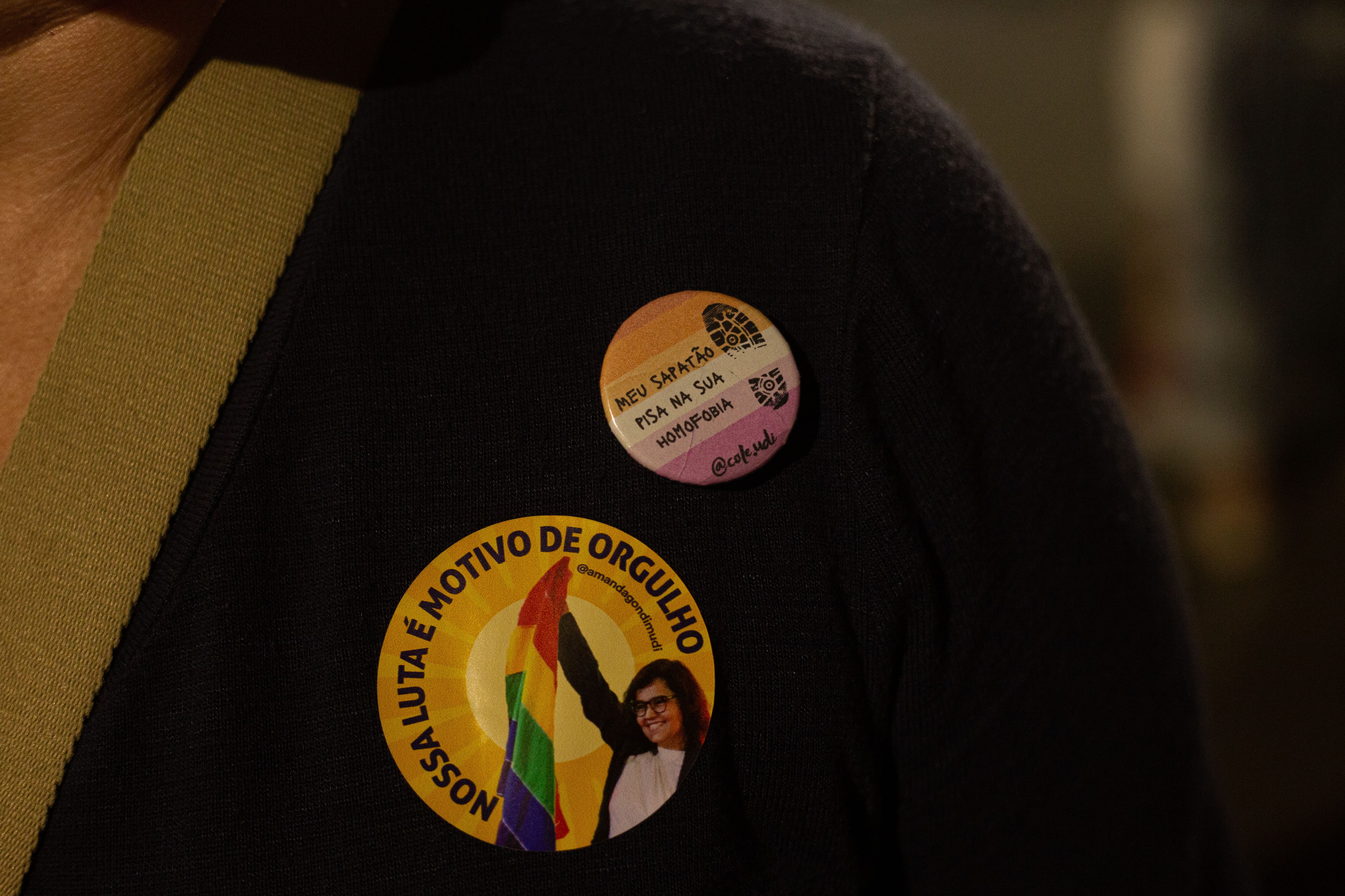 Foto em primeiro plano mostra blusa com adesivo da vereadora Amanda Gondim, segurando bandeira LGBTQIAPN+ e boton com a escrita 'Meu sapatão pisa na sua homofobia'
