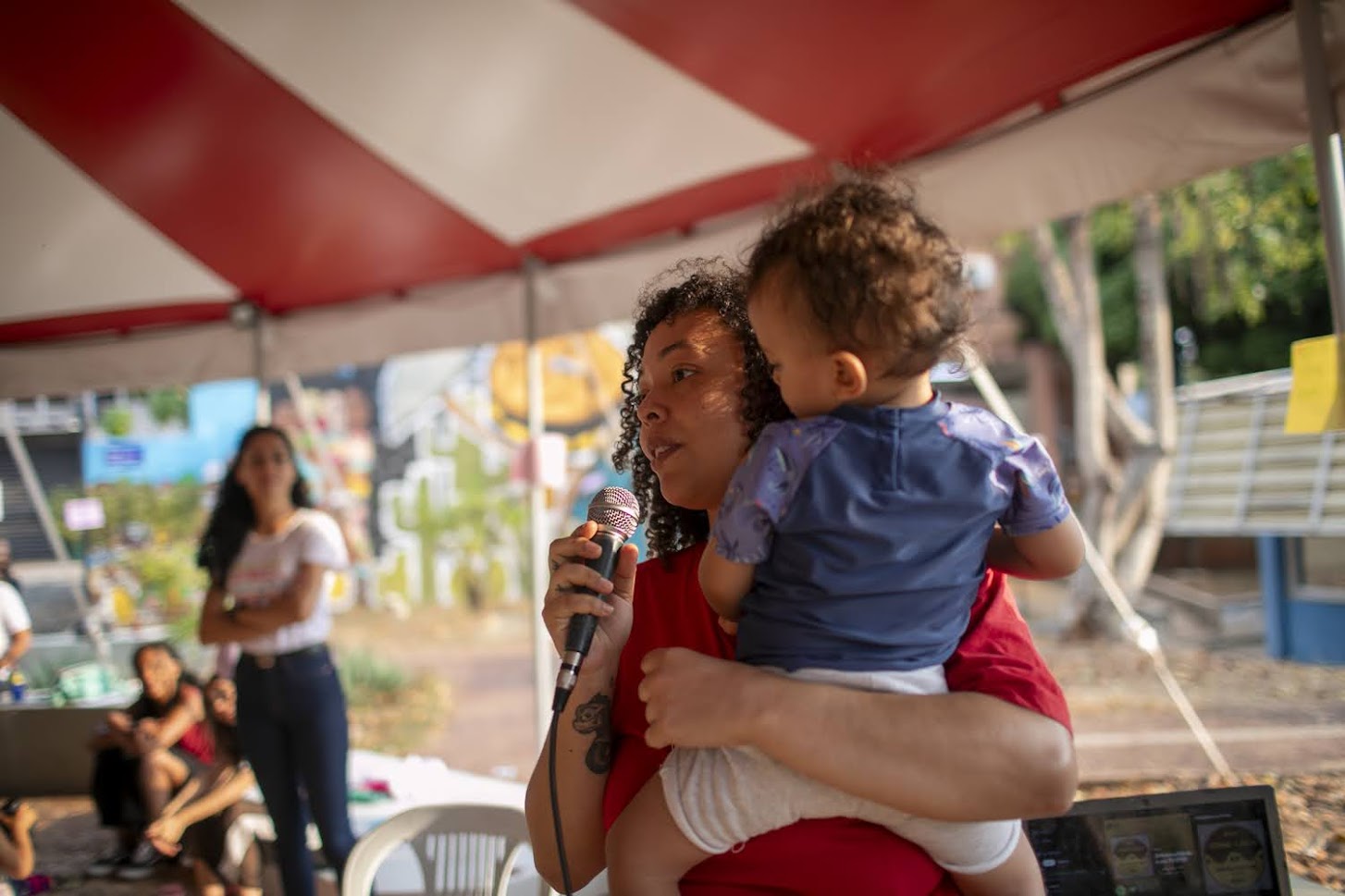 Imagem de Lays Braga falando ao microfone, segurando seu filho com um dos braços