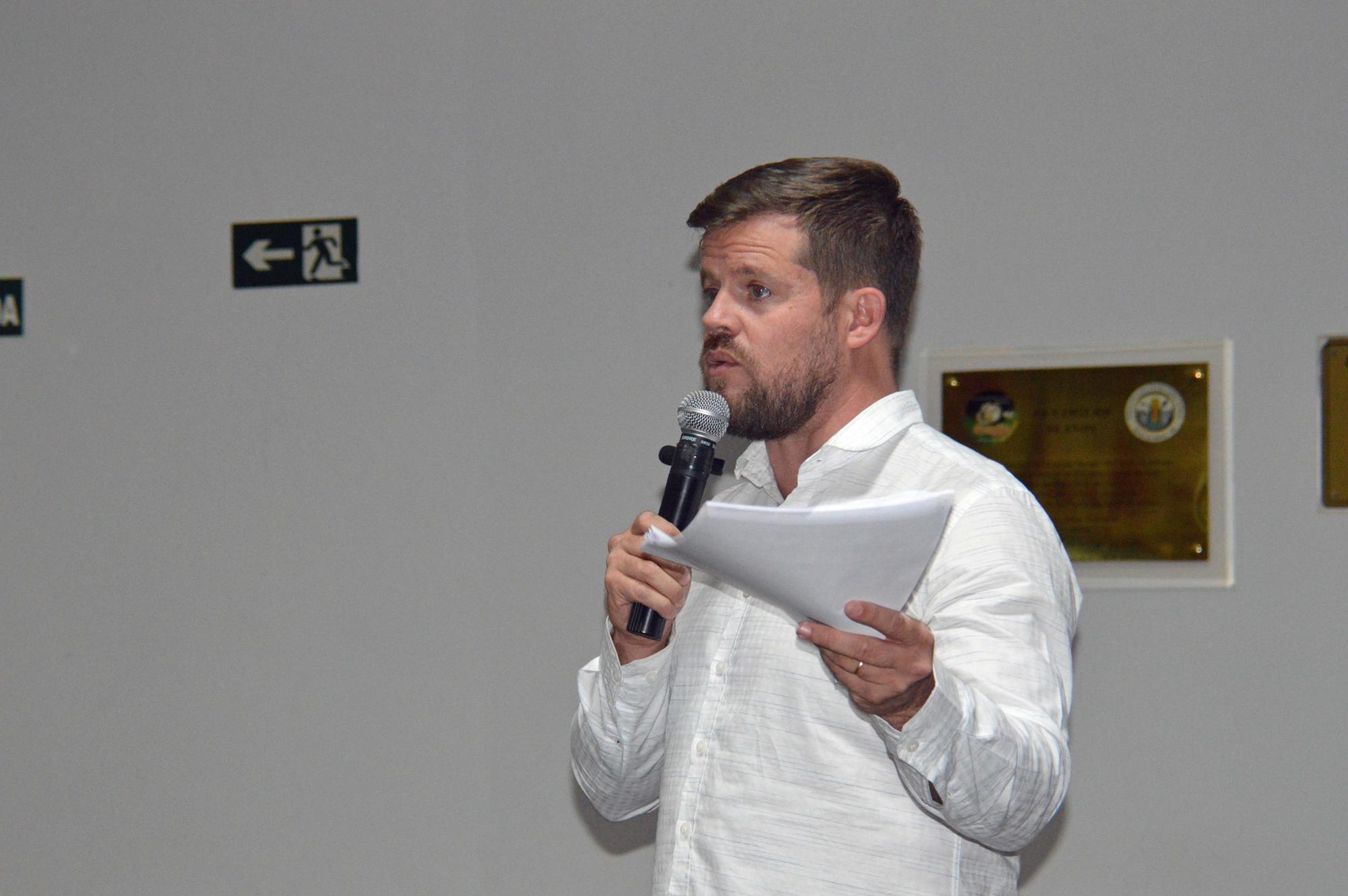 Homem branco, com barba, usando camisa social branca e segurando o microfone com a mão direita e folhas de papel na mão esquerda