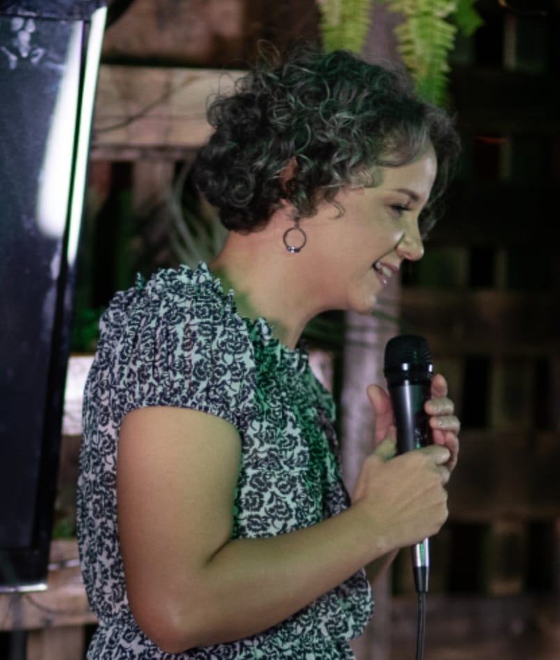 Imagem da professora Luciana Oliveira, de perfil, segurando um microfone e cantando
