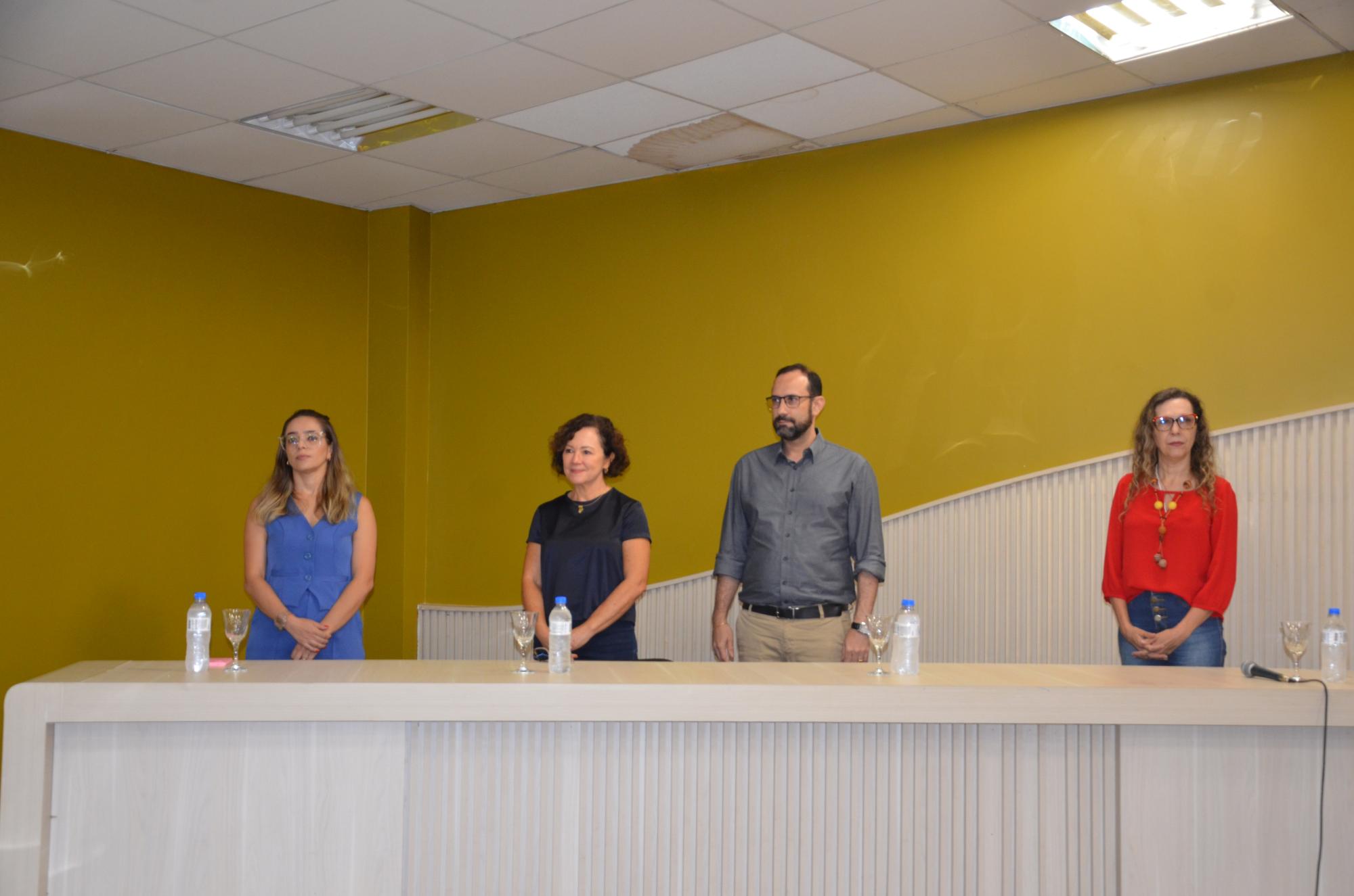 Na imagem, três mulheres e um homem, todos em pé e atrás de uma mesa na cor branca