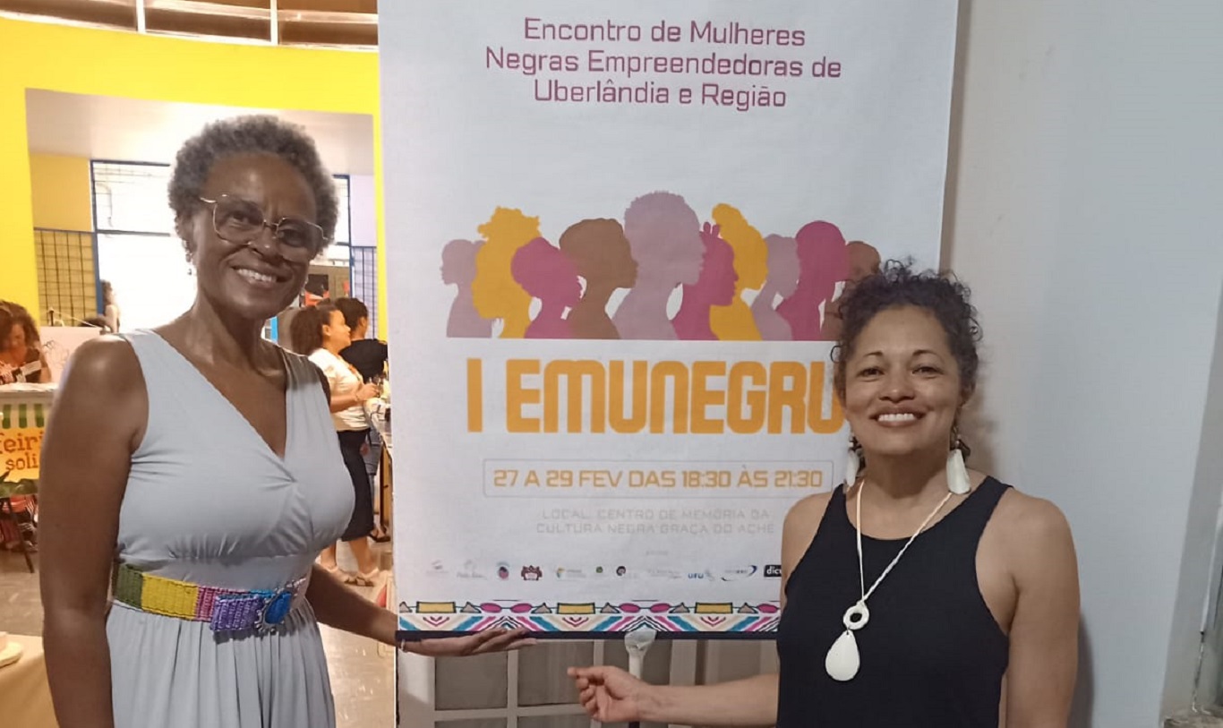 Na imagem, duas mulheres negras em pé, ao lado de um banner