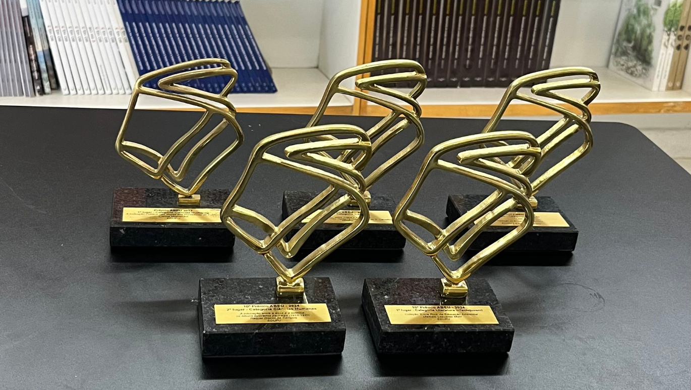 Imagem de cinco troféus expostos, em frente a uma prateleira com livros