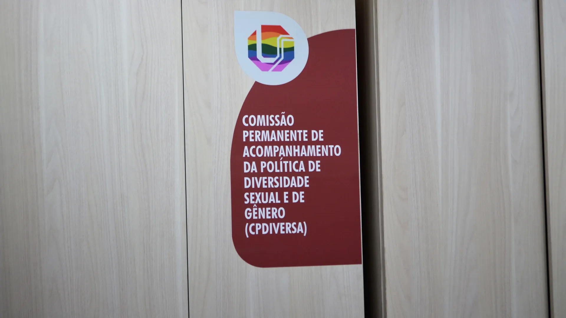 A imagem mostra uma placa sobre madeira clara com o logotipo da UF (Universidade Federal) em cores do arco-íris. Abaixo, está escrito: “Comissão Permanente de Acompanhamento da Política de Diversidade Sexual e de Gênero (CPDiversa)”.
