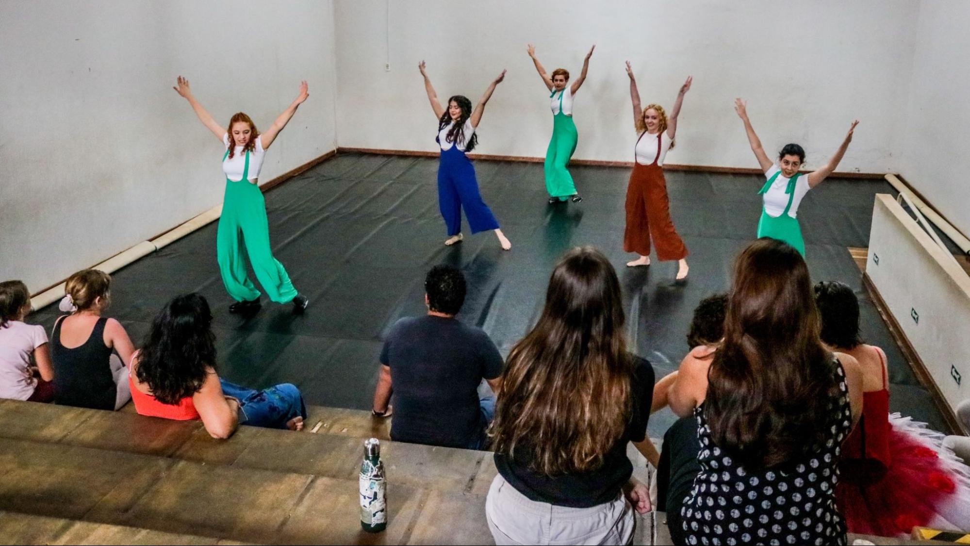 Pessoas vestidas com cores verdes, vermelhas e azuis, dançando em frente a outras pessoas que observam
