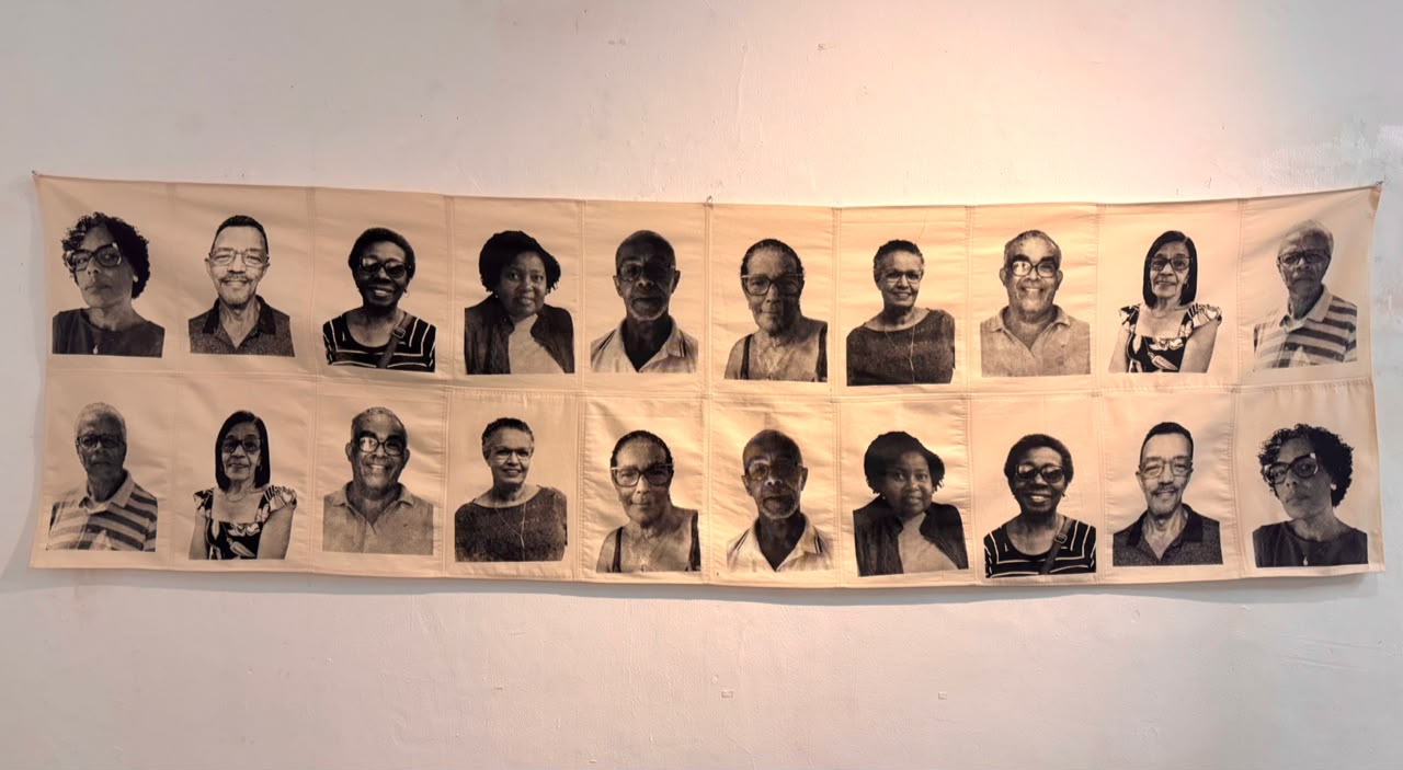 Obra têxtil composta por fotografias em preto e branco de diferentes pessoas negras costuradas sobre tecido claro. Os retratos estão organizados em duas fileiras horizontais e apresentam homens e mulheres de diferentes idades, com expressões serenas e sorridentes. A peça está exposta em uma parede branca e destaca temas relacionados à memória, ancestralidade e trajetórias familiares.