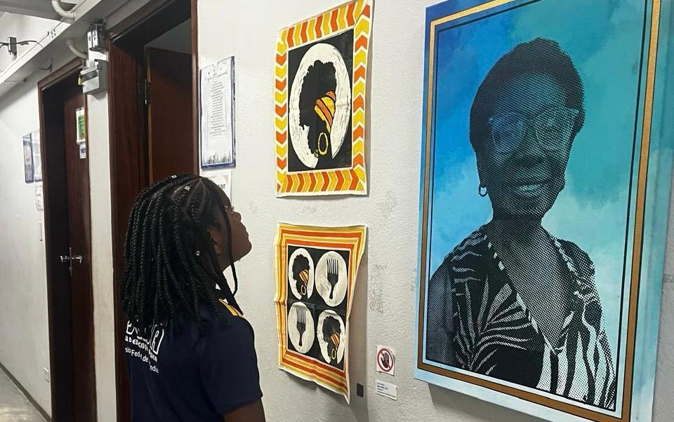 Estudante observa obras da exposição Costurando Memórias instaladas em um corredor do CAP/UFU. Na parede, há retratos e composições artísticas inspiradas em pessoas negras e em elementos da cultura afro-brasileira. Em destaque, uma grande obra em tons de azul apresenta o retrato de uma mulher idosa. A estudante está de perfil, observando as obras expostas.