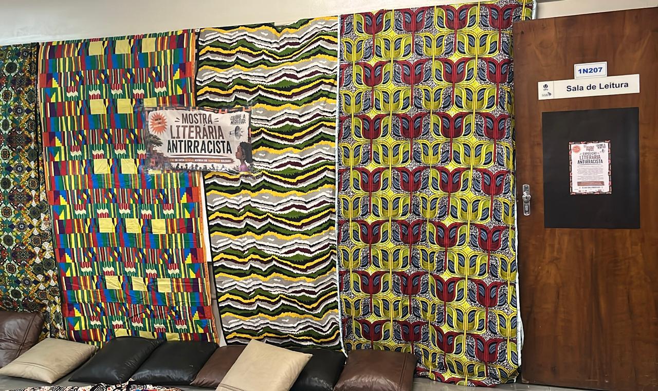 Entrada da sala de leitura decorada parapa a mostra Literária Antirracista. Tecidos estampados com padrões coloridos inspirados em estampas africanas revestem a parede ao lado da porta. Um cartaz anuncia a exposição, enquanto almofadas dispostas no chão criam um espaço acolhedor para leitura, convivência e atividades relacionadas à valorização da literatura e da cultura afro-brasileira.