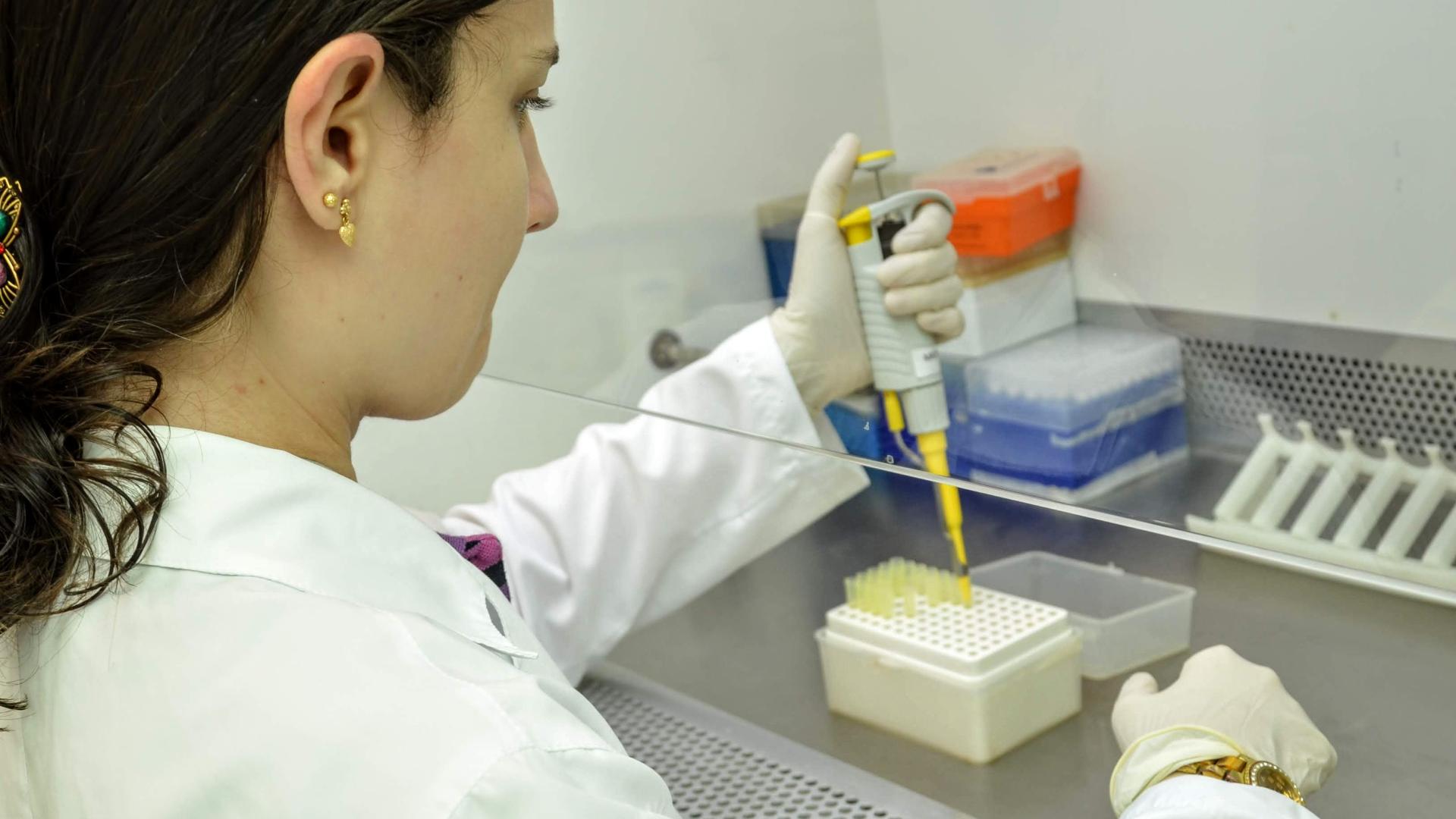 Foto colorida com mulher de costas vestida com jaleco branco, trabalhando em laboratório