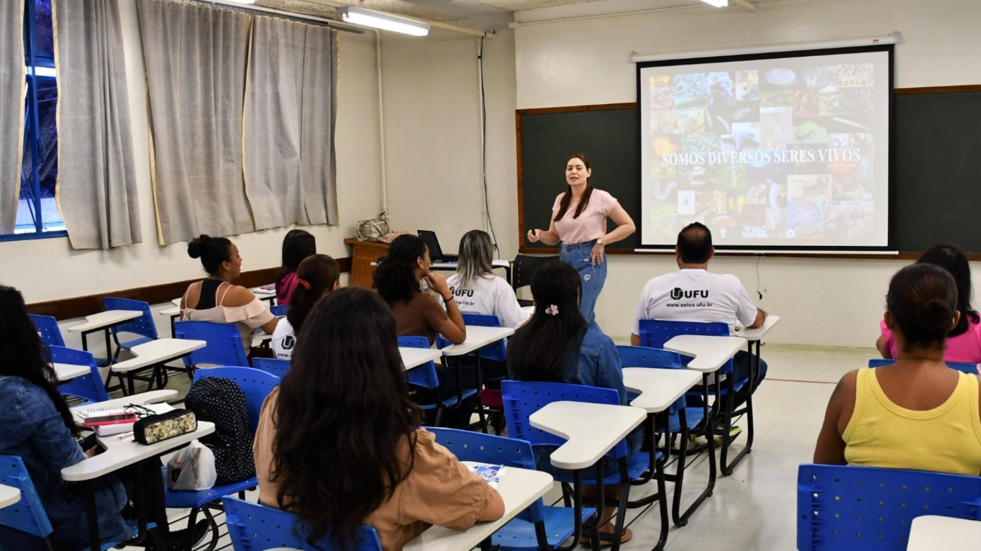 Imagem de uma professora em pé, ministrando aula, diante de um grupo de alunos de costas, sentados em suas carteiras numa sala da Escola Técnica de Saúde da UFU