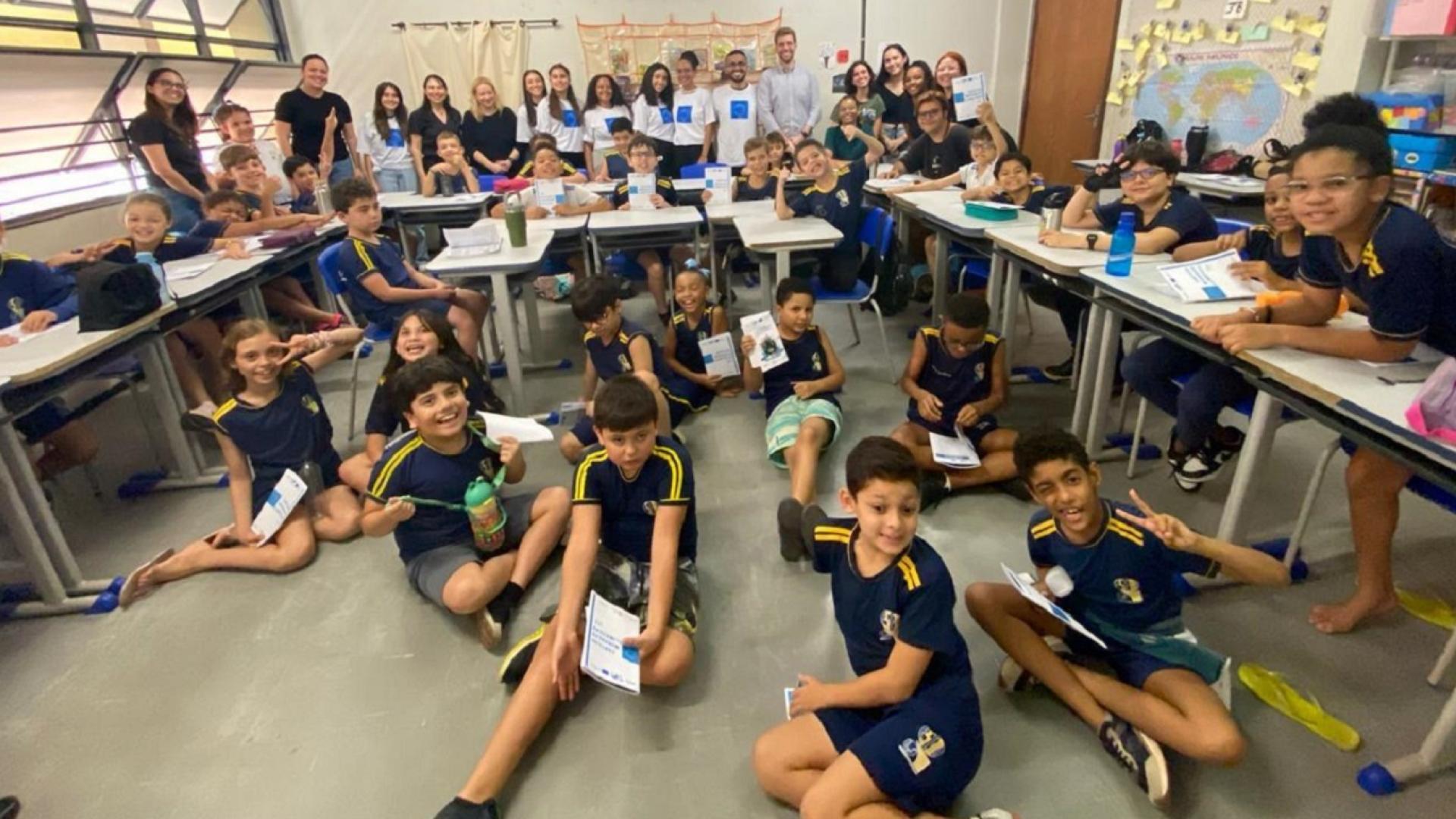 Grupo de crianças em uma sala de aula; algumas estão sentadas no chão e outras em suas carteiras; ao fundo, adultos em pé