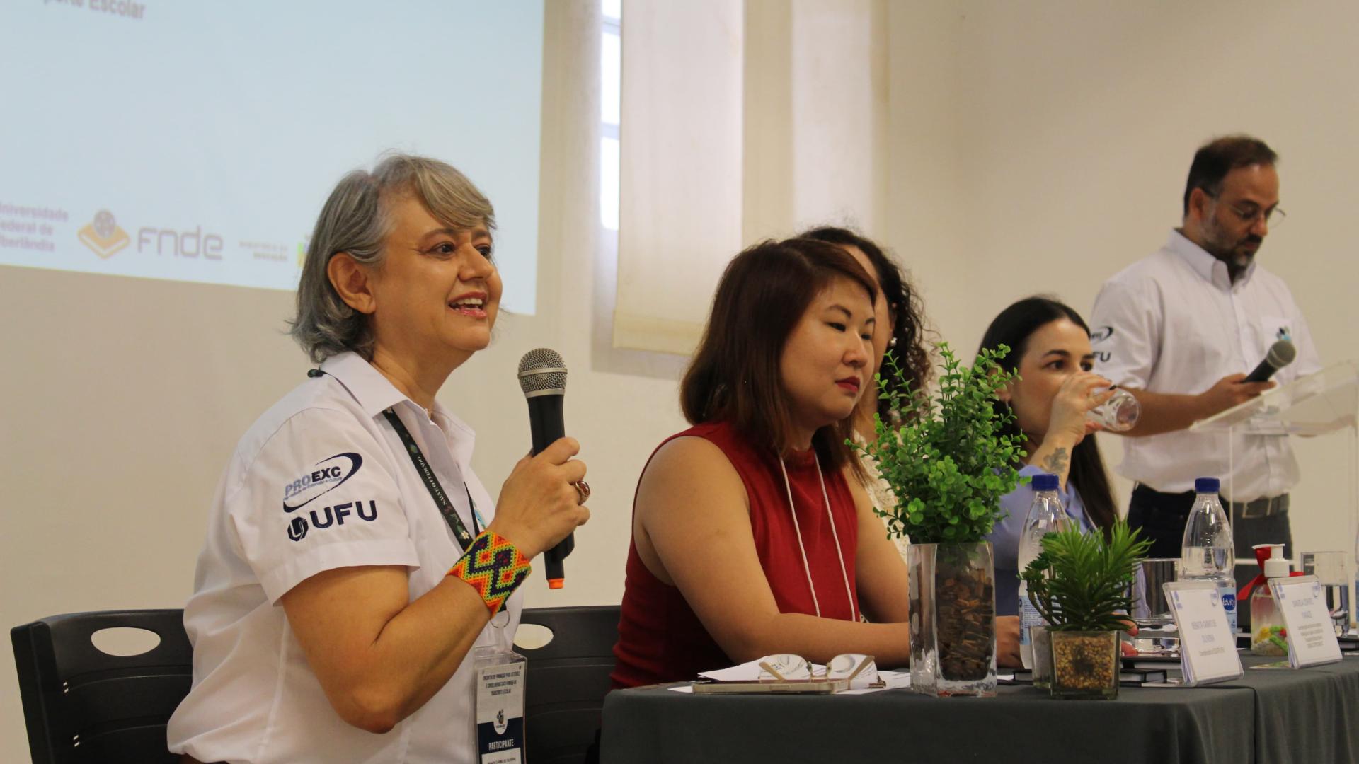 Quatro mulheres sentadas, perfiladas na mesa de abertura de um evento; a mais à esquerda fala ao microfone