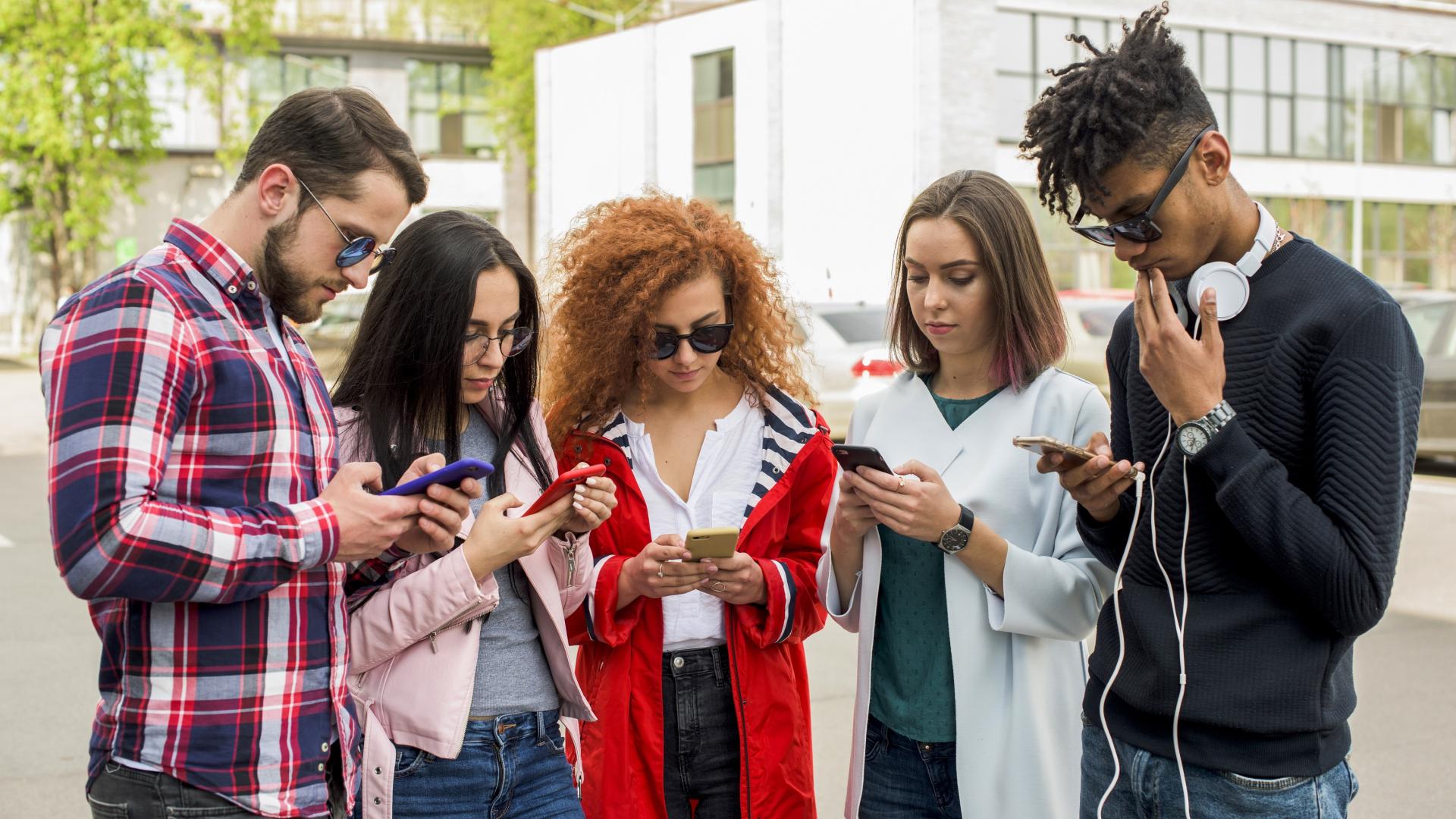 Imagem de banco de imagens. Cinco jovens adultos, sendo três mulheres e dois homens; todos mexendo no celular