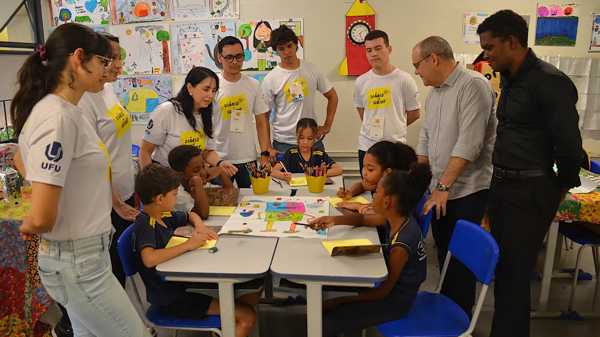 Registro de um grupo de 8 adultos em pé, observando 5 crianças sentadas, fazendo atividades escolares