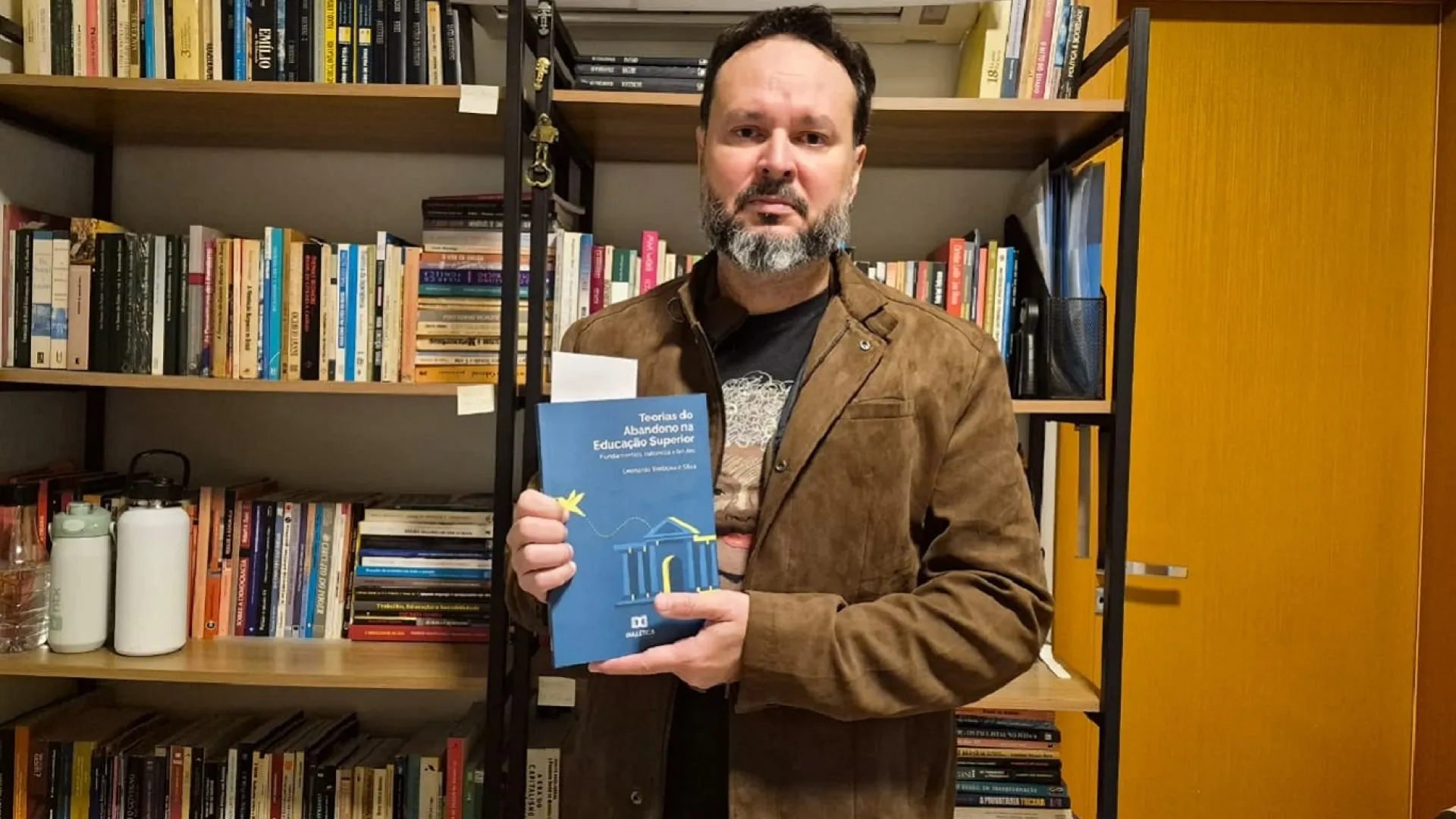 Professor Leonardo Barbosa e Silva posa, exibindo o seu livro, à frente de uma estante onde estão muitas outras obras literárias