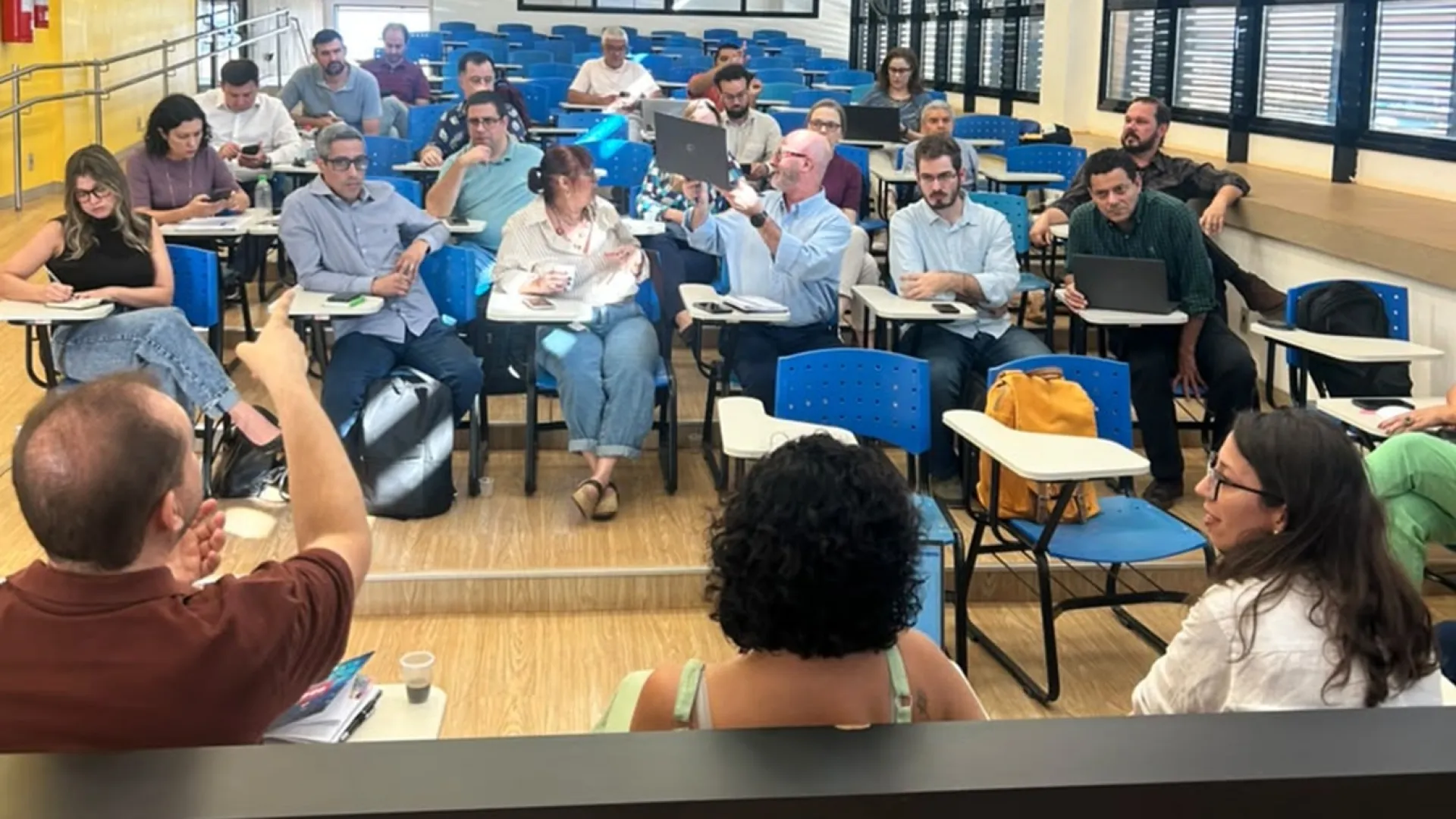 Foto com três pessoas de costas, sendo um homem (à esquerda) gesticulando e duas mulheres; à frente, está um grupo de 20 pessoas