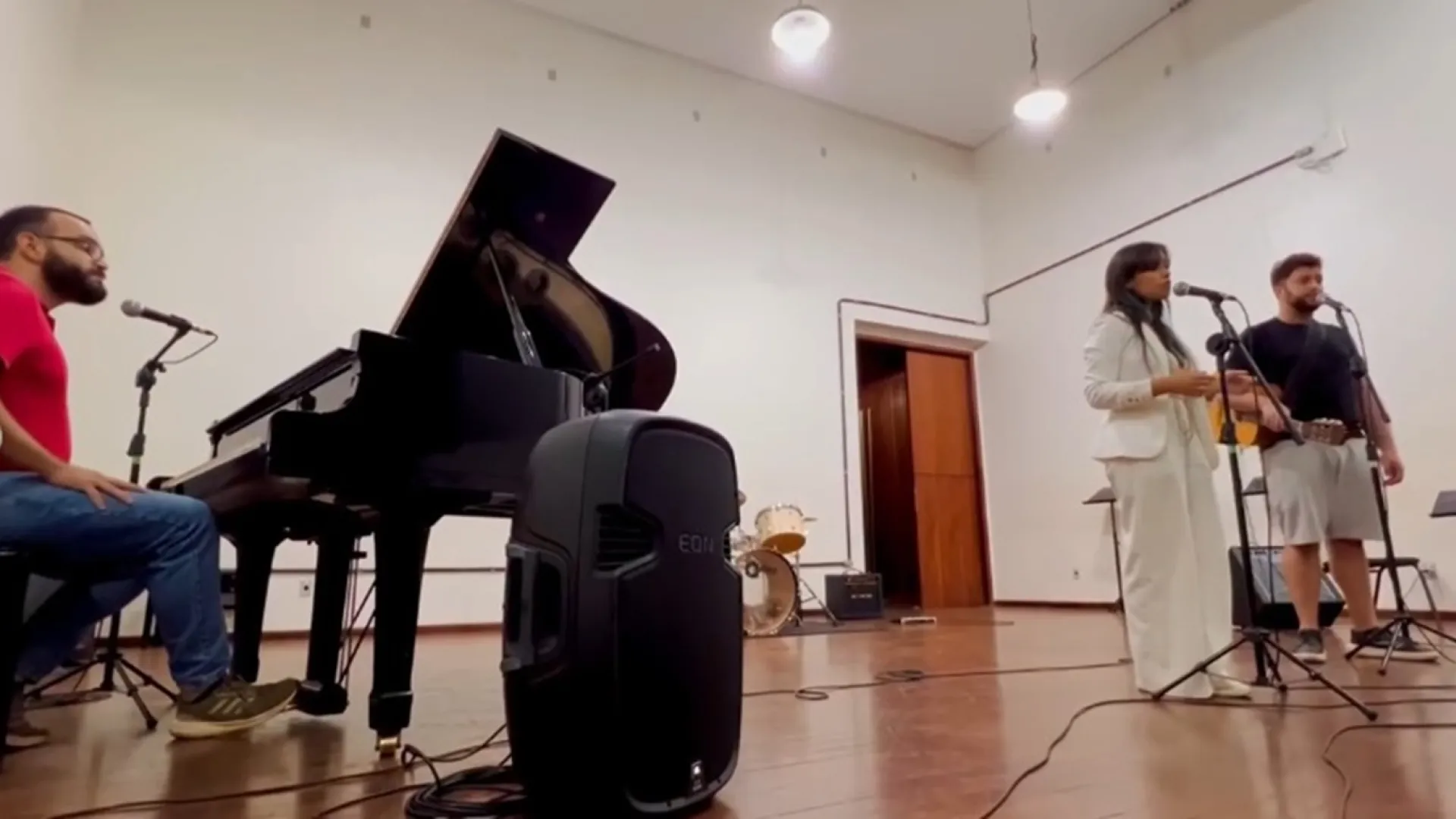 Imagem de apresentação musical, com um homem sentado ao piano (à esquerda) e uma moça e um rapaz em pé, diante de microfones (à direita)