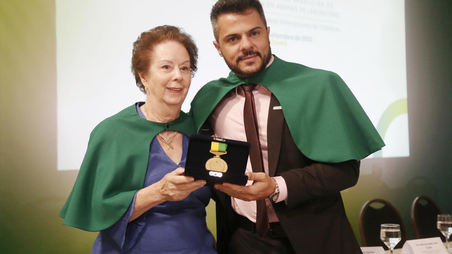 Foto colorida traz Ekaterina, patrona da cadeira 1, entregando a medalha de acadêmico para Murilo, patrono da cadeira 4