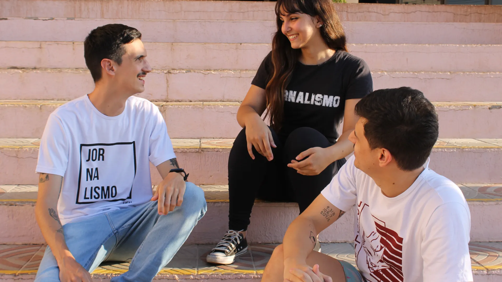 Três estudantes de Jornalismo conversando sentados numa escada, sendo uma mulher e dois homens