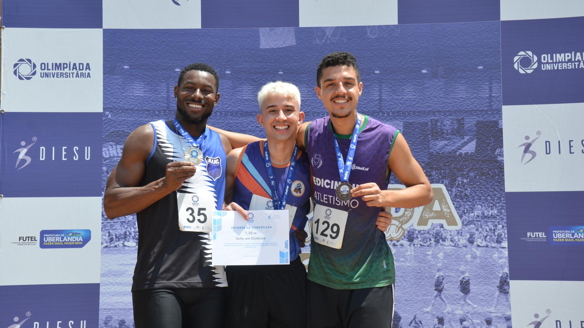 Três estudantes-atletas do sexo masculino no pódio com suas respectivas medalhas