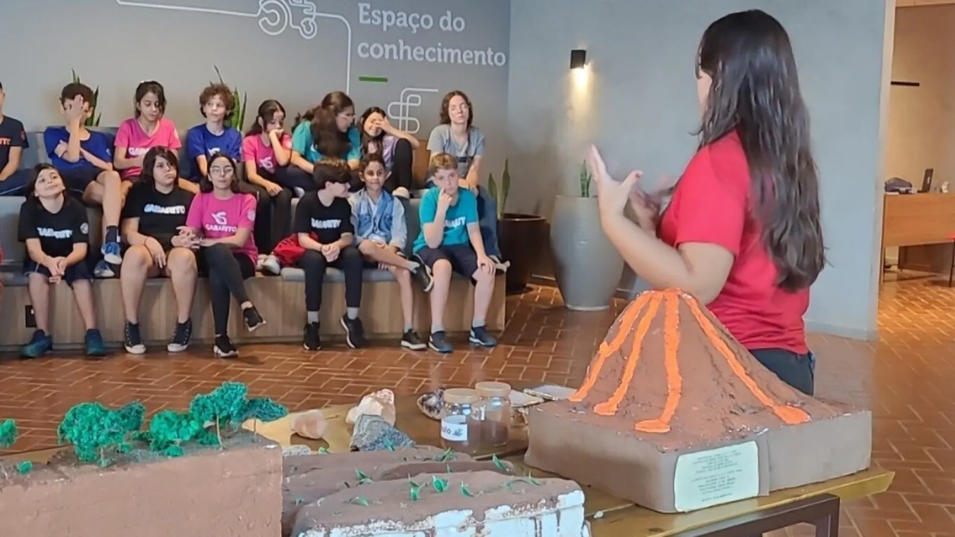 Imagem de uma bolsista do projeto mostrando uma maquete que explica sobre o solo e os vulcões. Na plateia, há jovens observando a apresentação