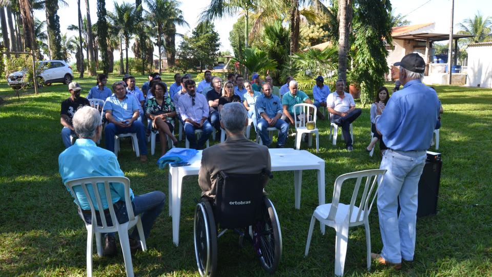 Na imagem, há dois homens falando à frente de uma pequena plateia