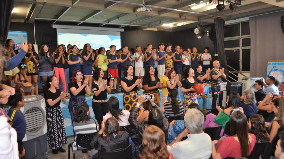 Um grupo de crianças e mulheres canta enquanto uma plateia assiste