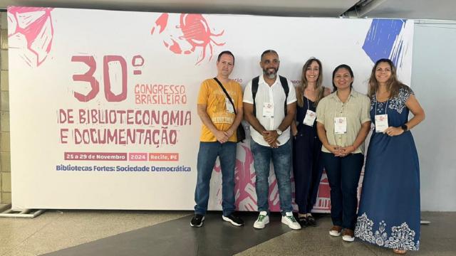 Cinco pessoas posando em frente a um painel com o nome do evento; são dois homens e três mulheres