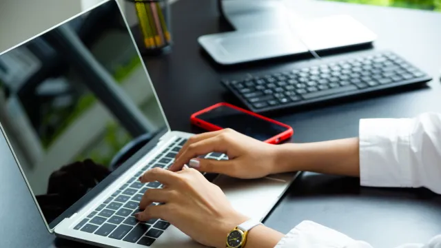 Mãos digitando em um notebook, com celular ao lado