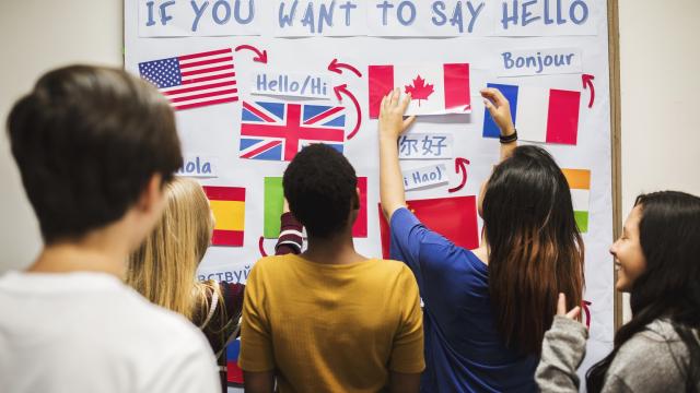 Jovens diante de um mural com bandeiras de vários países e as inscrições de como é dito 'olá' nos idiomas neles falados