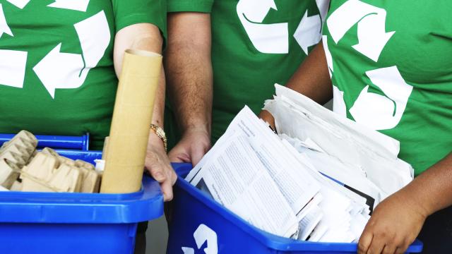 Três pessoas segurando caixotes azuis de plástico, cheios de materiais recicláveis. Elas estão vestidas em camisetas verdes, estampadas com o símbolo da reciclagem em branco.