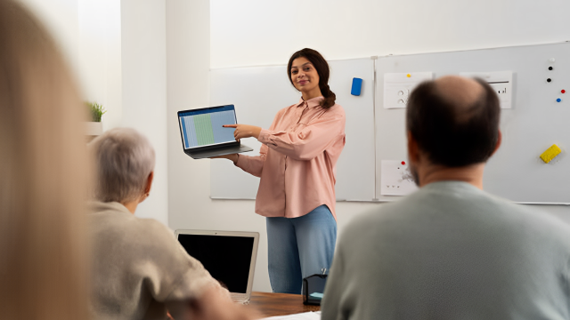 Imagem de uma professora em pé ministrando aula, mostrando a tela de um notebook; em primeiro plano, é possível ver sentados, de costas, uma mulher de cabelo grisalho e um homem calvo