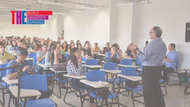 Foto mostra o diretor de Relações Internacionais da UFU, Waldenor Barros Moraes Filho, em pé e falando ao microfone, numa sala de aula, diante de um grupo de estudantes sentados em carteiras azuis