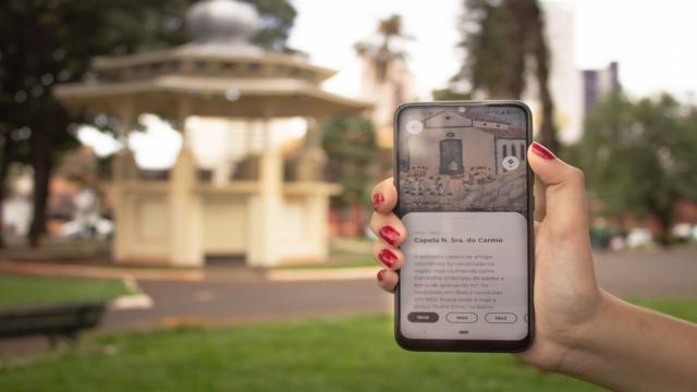 Atrás da mão que segura o celular, é possível ver o coreto da Praça Clarimundo Carneiro, enquanto o aplicativo criado é exibido na tela do dispositivo