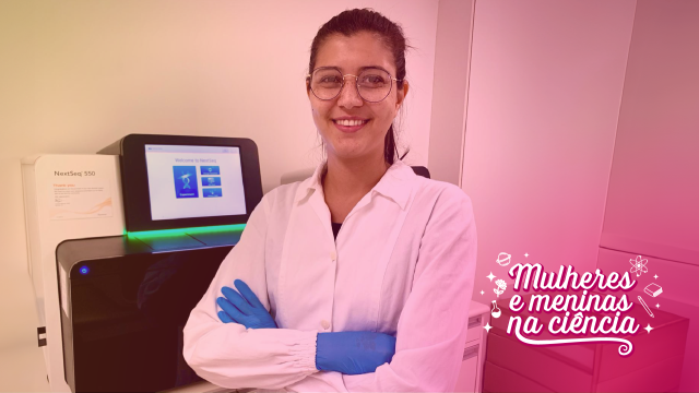Mulher pesquisadora em laboratório está em pé, com jaleco branco, braços cruzados e sorrindo