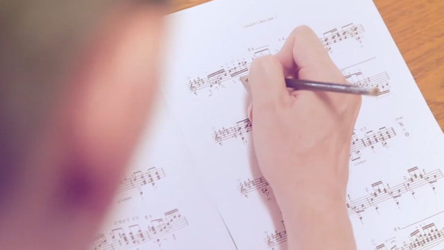 Imagem com um rosto desfocado, em primeiro plano; ao fundo, uma mão segurando um lápis e escrevendo em uma partitura  