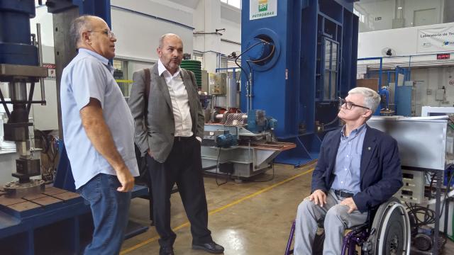 Três homens, sendo um deles cadeirante, conversando em um laboratório de engenharia mecânica