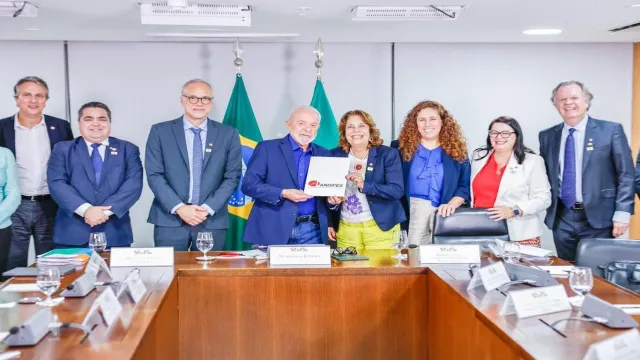 Imagem mostra grupo de pessoas em pé, reunidas, em volta de uma grande mesa. As duas pessoas posicionadas no meio da foto seguram um documento com a escrita 'Andifes.